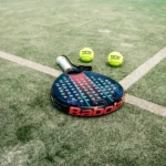 A blue and black Babolat padel racket lies on a grass court with three yellow padel balls nearby, each marked 