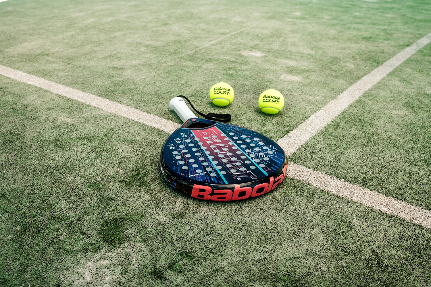 A blue and black Babolat padel racket lies on a grass court with three yellow padel balls nearby, each marked 