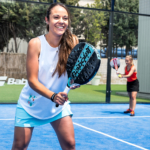 A woman in a white tank top and light blue shorts is playing padel on a blue court, holding a padel racket and smiling. Demonstrating how to play padel, she enjoys the game as another player in the background, wearing a red shirt and black skirt, also holds a racket. Trees and a fence are visible in the background. © Powerleague