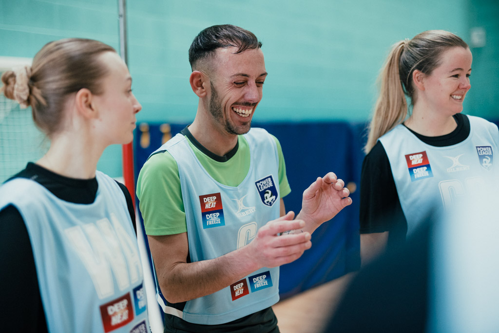 Three people, wearing sports bibs with sponsorship logos, smile and laugh together, standing in what appears to be an indoor sports venue with a net visible in the background. © Powerleague