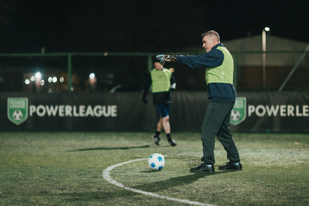A person wearing a green bib and gloves is on an outdoor football field at night, playing as a goalkeeper. There's a blue and white football in front of them. Another player is in the background. The field is surrounded by a fence with 