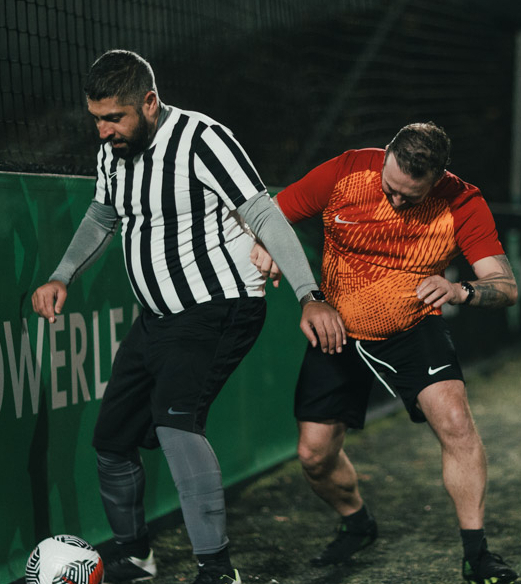 Two men are playing soccer on an outdoor field. The man on the left, in a black-and-white striped shirt, controls the ball while closely guarded by the man on the right, wearing a red-orange shirt. It's nighttime, with a dark fence and green advertisement board in the background. © Powerleague