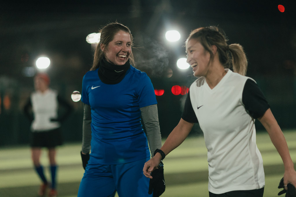 Two women, one in a blue soccer uniform and the other in a white and black uniform, are smiling and walking together on a lit outdoor soccer field at night. Both are wearing gloves, and there are other players and lights visible in the background. © Powerleague