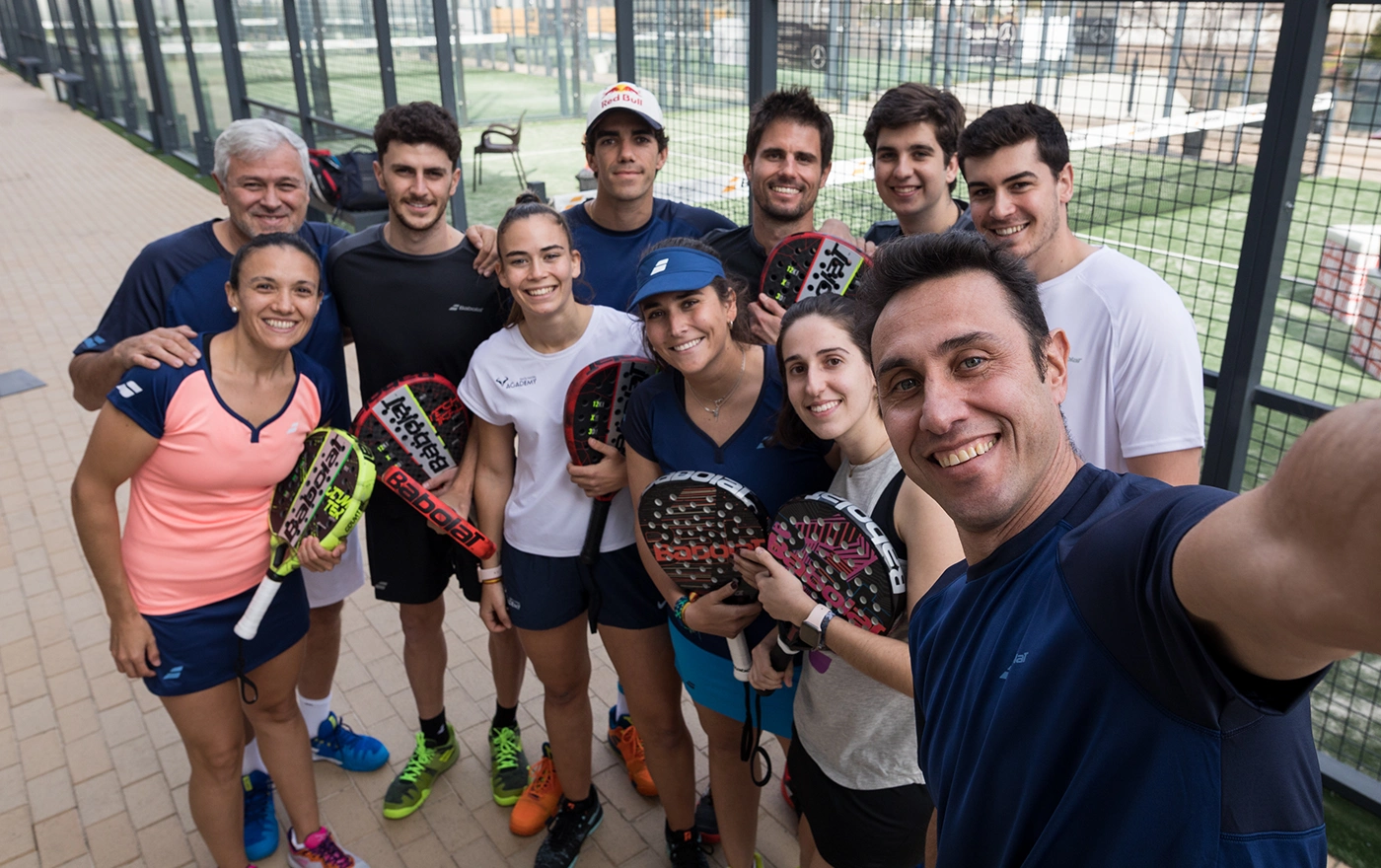 A group of eleven people is gathered for a selfie on a padel tennis court. They are smiling, dressed in athletic clothing, and holding paddles. The court is surrounded by a fence with glimpses of other courts and buildings in the background. © Powerleague