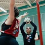 Two local netball team players are in action on an indoor court. The player in the foreground, wearing a black jersey, is attempting a shot with the ball raised overhead. The player in the background, wearing a red bib, is defending with an outstretched arm. © Powerleague