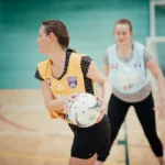 Two women from local netball teams are playing indoors. One player in a yellow bib holds a netball, preparing to pass, while a defender in a blue bib reaches out to block. The players are on a wooden court with a netball goalpost in the background. © Powerleague