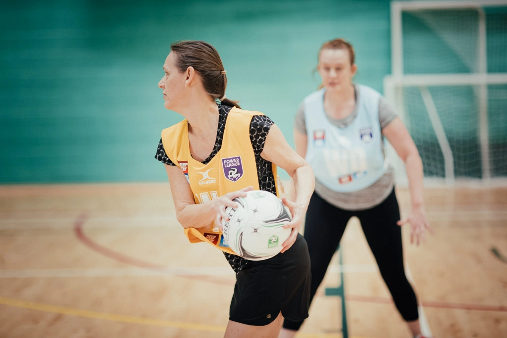 Two women from local netball teams are playing indoors. One player in a yellow bib holds a netball, preparing to pass, while a defender in a blue bib reaches out to block. The players are on a wooden court with a netball goalpost in the background. © Powerleague