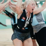 A woman in a sports uniform excitedly holds a trophy above her head while running through a tunnel made by her mixed-netball teammates' raised arms in a gymnasium. She wears a medal around her neck and has a joyful facial expression. © Powerleague