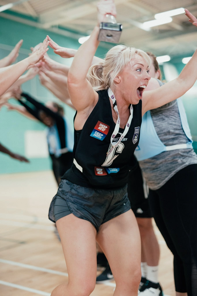 A woman in a sports uniform excitedly holds a trophy above her head while running through a tunnel made by her mixed-netball teammates' raised arms in a gymnasium. She wears a medal around her neck and has a joyful facial expression. © Powerleague