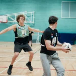 Two male athletes are playing mixed-netball indoors. The player in the foreground, wearing a black shirt, holds the ball while the player in the background, wearing a green bib, extends his arms to block. Both are in motion on a wooden court with a basketball hoop in the background. © Powerleague