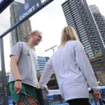 Two people are seen entering a fenced padel court marked 