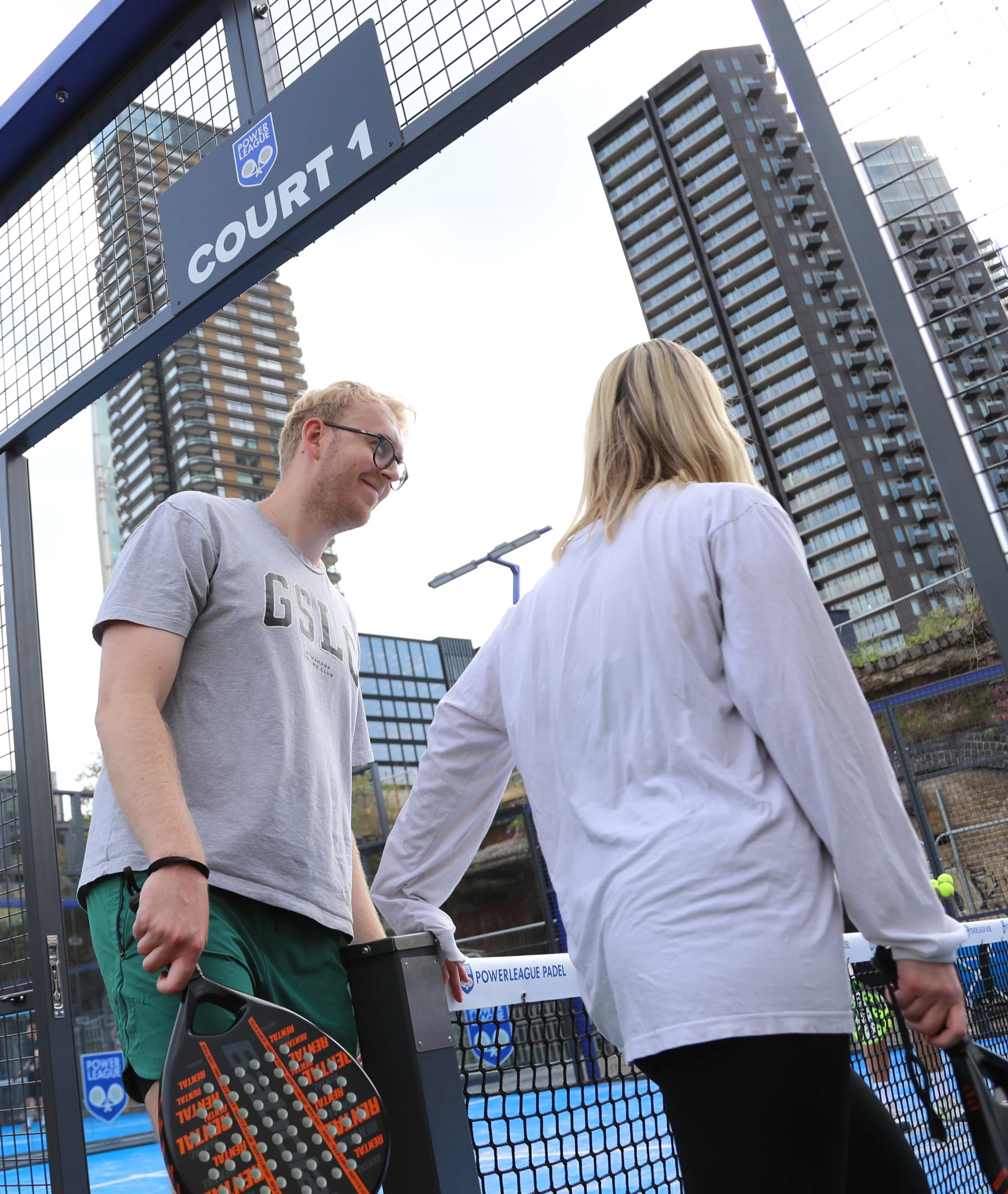 Two people are seen entering a fenced padel court marked 