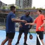 Three men on a blue-court padel court are engaging in a light-hearted moment. Two of them, in dark athletic wear, smile and extend hands toward a third player in an orange shirt. They stand near the net with modern city buildings in the background. © Powerleague