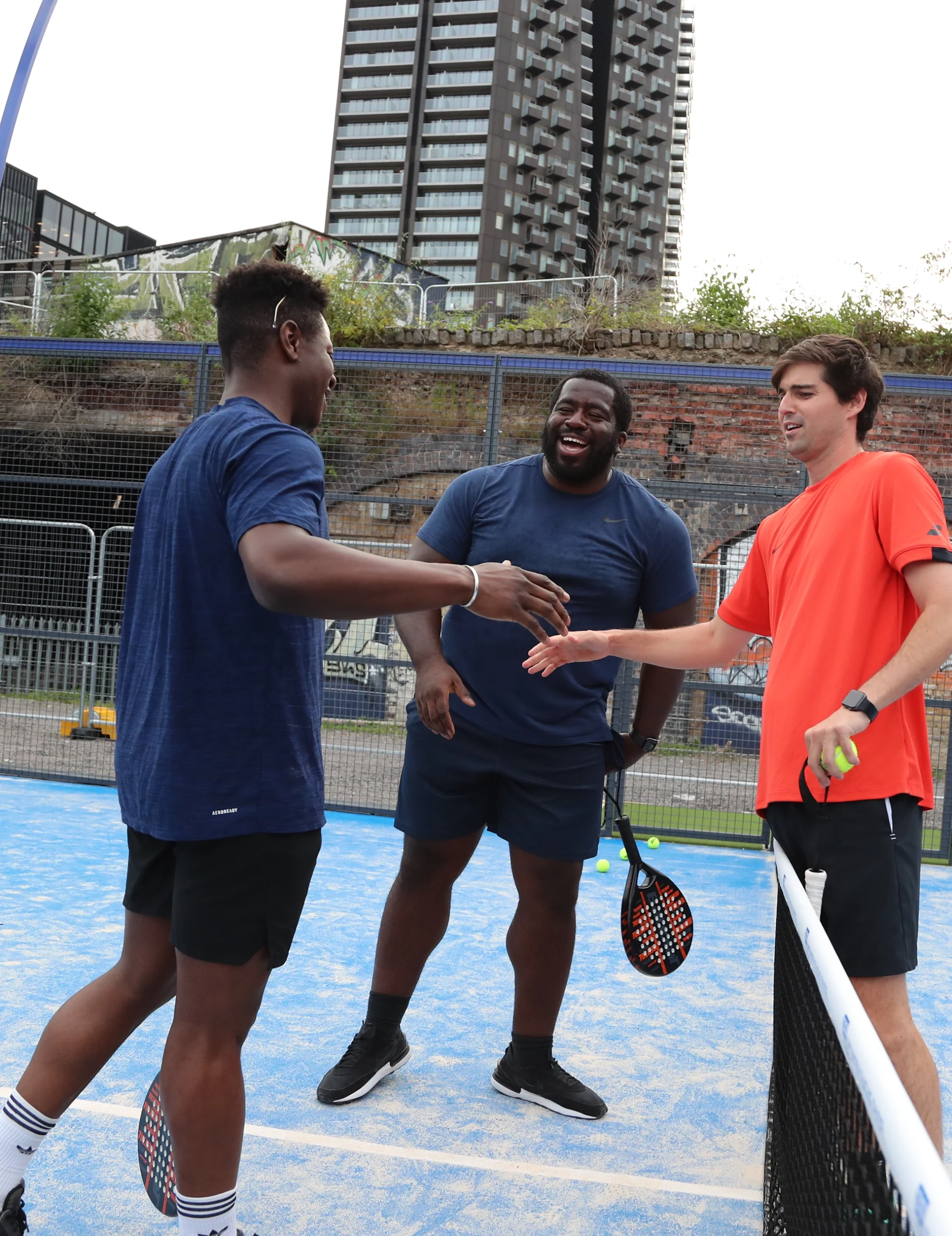 Three men on a blue-court padel court are engaging in a light-hearted moment. Two of them, in dark athletic wear, smile and extend hands toward a third player in an orange shirt. They stand near the net with modern city buildings in the background. © Powerleague