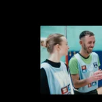 Three individuals wearing sports bibs are smiling and conversing indoors, seemingly in a mixed-netball gym or sports hall. They stand near a net, with the person in the center laughing while holding something in their hands. The overall mood is cheerful and lighthearted. © Powerleague