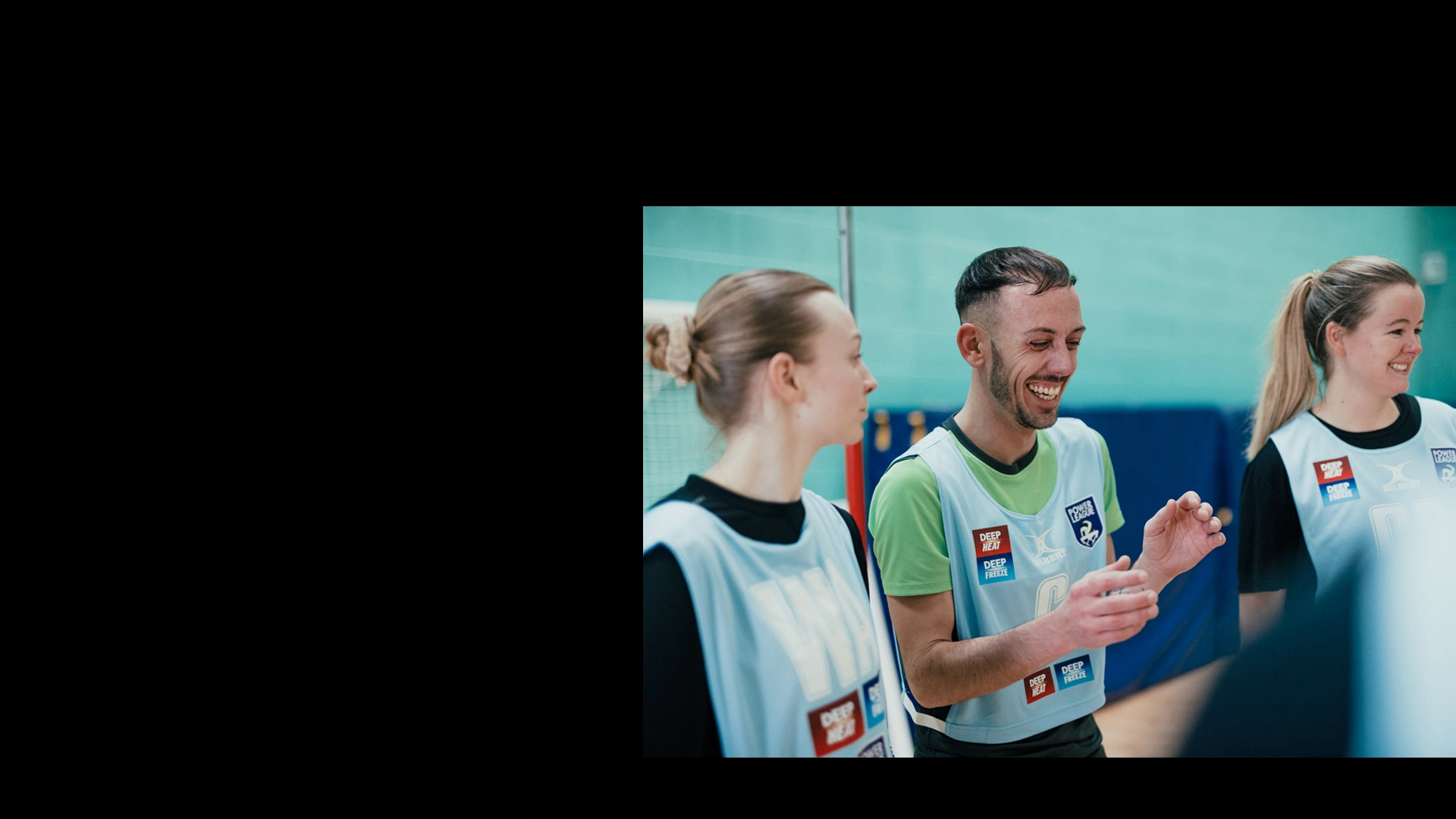 Three individuals wearing sports bibs are smiling and conversing indoors, seemingly in a mixed-netball gym or sports hall. They stand near a net, with the person in the center laughing while holding something in their hands. The overall mood is cheerful and lighthearted. © Powerleague