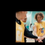 A group of Mixed-Netball players wearing yellow jerseys stand in a gymnasium with their hands stacked in the center, symbolizing team unity. The players are smiling and appear engaged, ready for their game or practice session. One player with curly hair stands out in the middle. © Powerleague