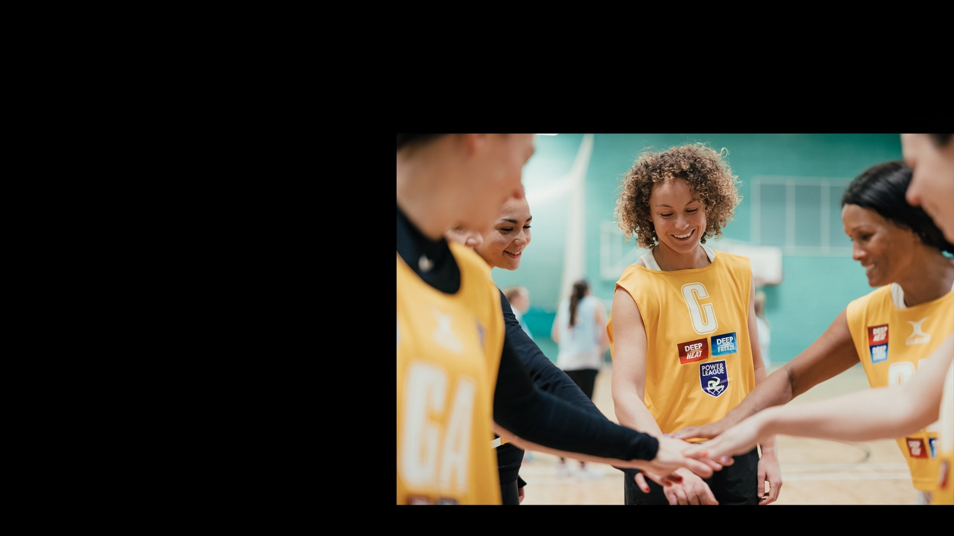 A group of Mixed-Netball players wearing yellow jerseys stand in a gymnasium with their hands stacked in the center, symbolizing team unity. The players are smiling and appear engaged, ready for their game or practice session. One player with curly hair stands out in the middle. © Powerleague