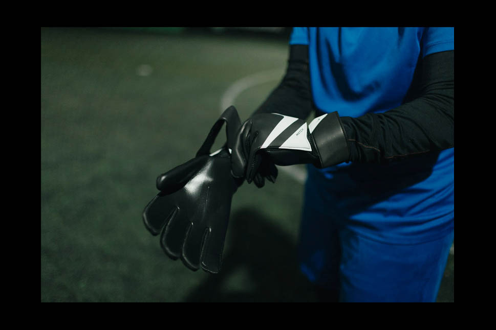 A soccer player wearing a blue uniform and black long sleeves is seen adjusting black gloves on a dimly lit field, almost like preparing for some legal stuff. Only the torso and arms are visible in the image. The background is out of focus. © Powerleague