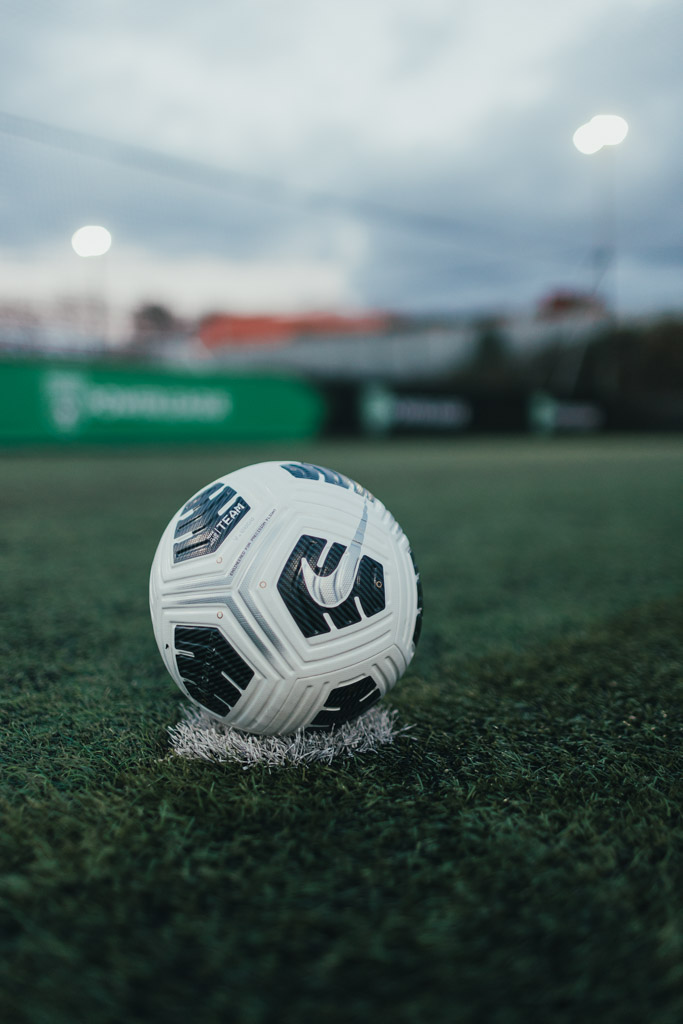 A close-up of a white and black soccer ball on a green artificial turf field, resting on a small patch of white marking. In the blurred background, there are lights on poles and a green banner, indicating an outdoor sports facility—perhaps where careers at Powerleague begin.