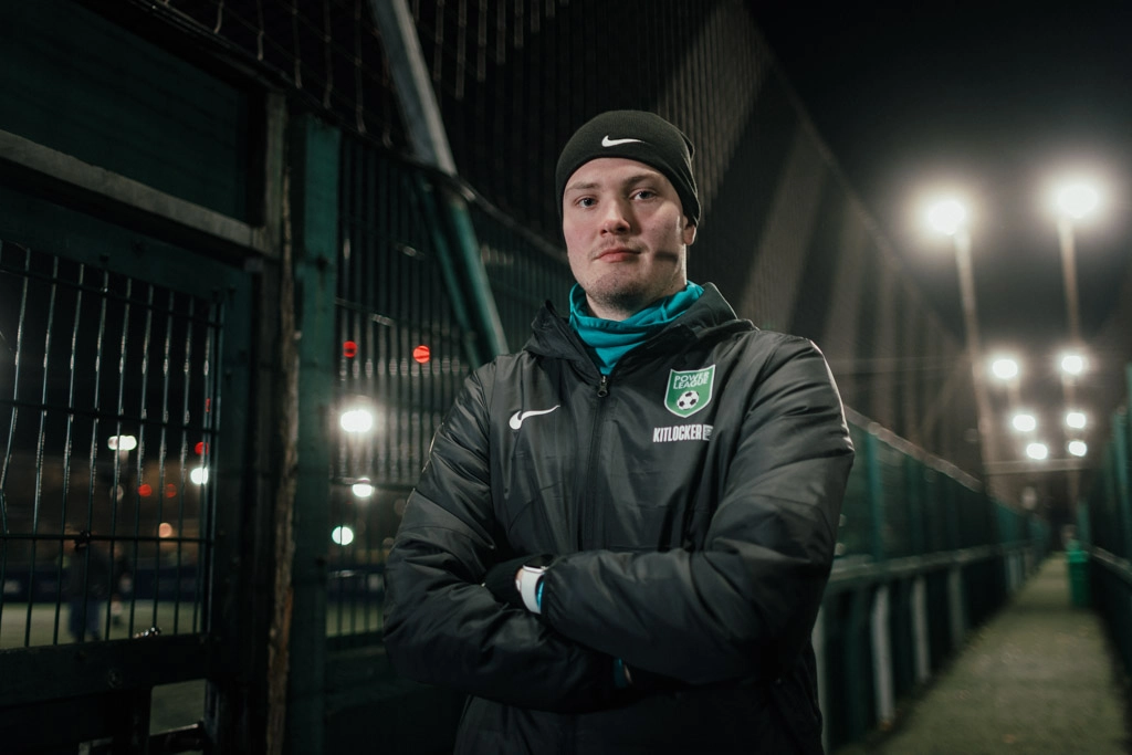 A person in a black beanie and jacket stands with arms crossed on a dimly lit soccer field. The individual has a focused expression and wears a green emblem with a soccer ball and 