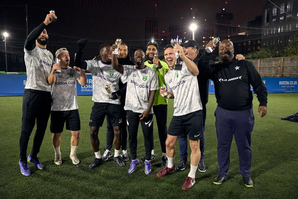 A group of men on an outdoor soccer field at night are celebrating, holding water bottles. They are wearing matching gray jerseys and shorts, and a coach dressed in black is standing with them. Buildings and field lights illuminate their 5-a-side football victory in the background. © Powerleague
