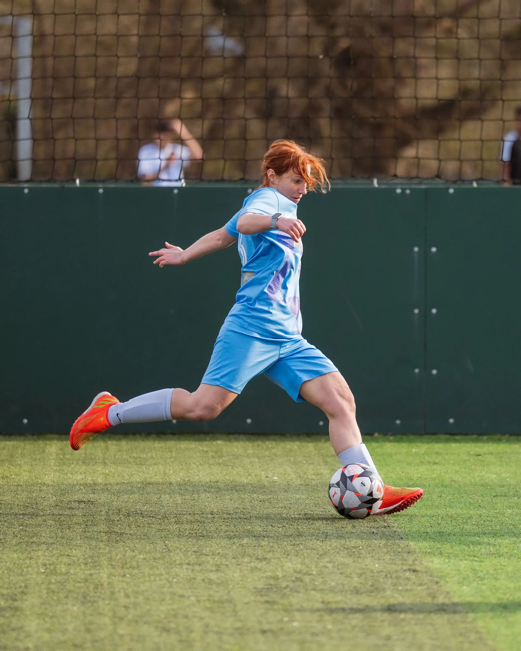 A women's football player in a light blue uniform and red cleats is about to kick a ball on a green field. The player's leg is extended back, and they appear focused on the ball. A dark green fence and spectators in the background are visible. © Powerleague