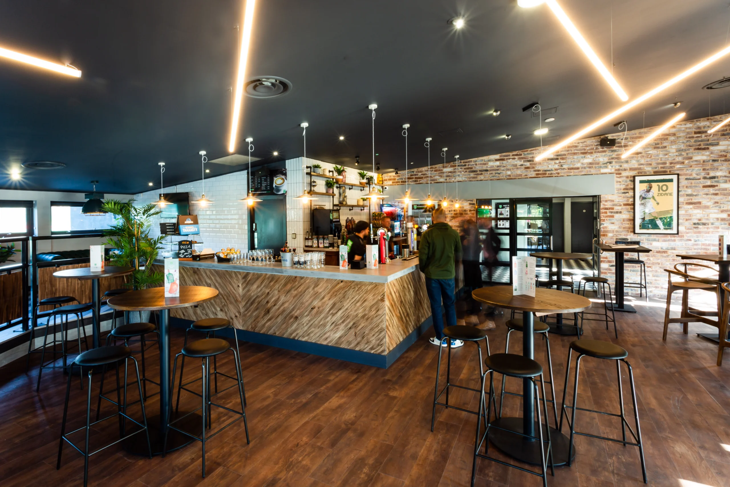 Modern bar interior with an industrial design featuring exposed brick walls and wooden accents. High tables and bar stools are arranged around a central bar counter. Pendant lights hang from the ceiling, and people are seen socializing in the background, much like careers at Powerleague. © Powerleague