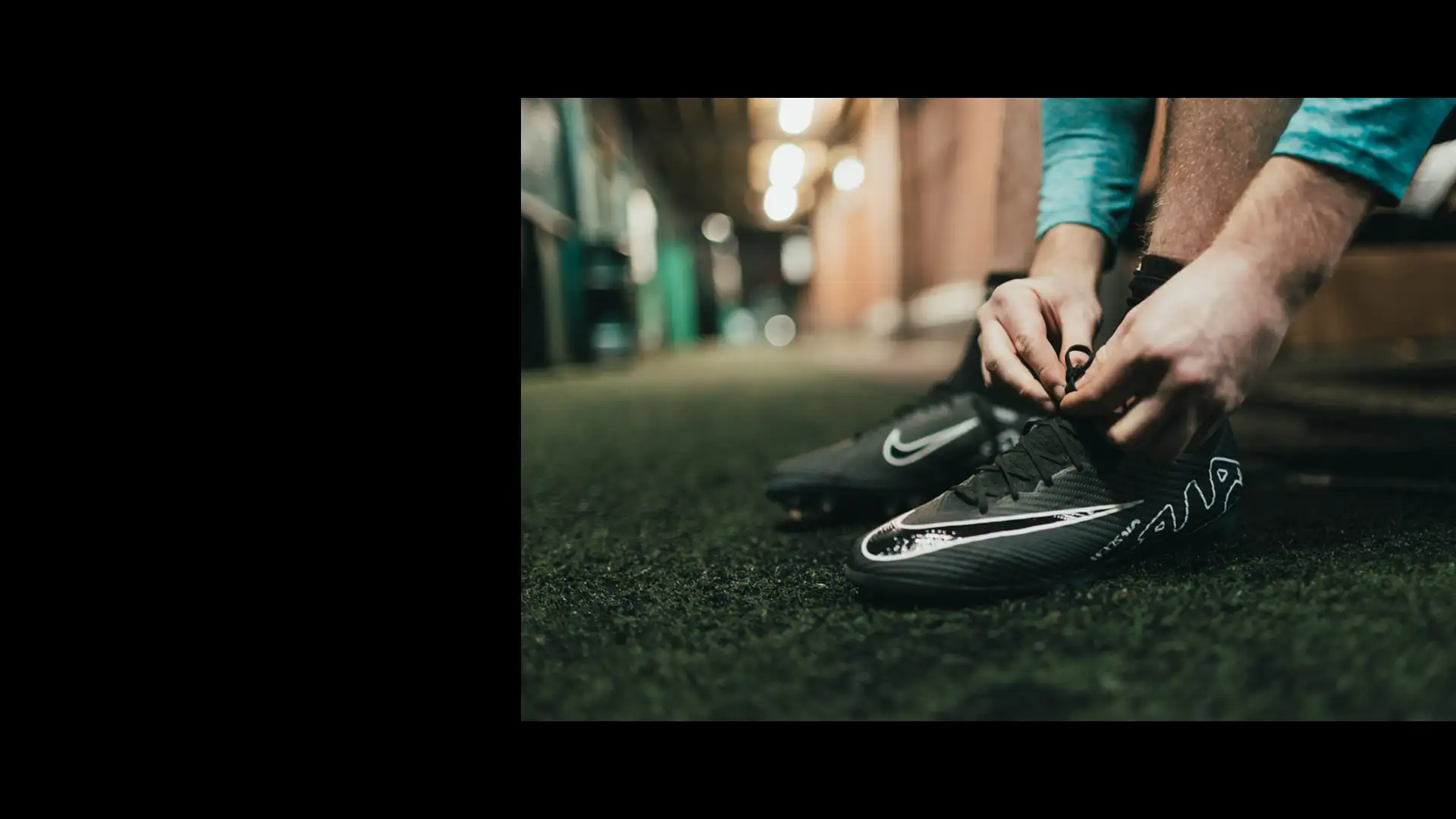 A person is tying the laces of their black Nike athletic shoes, preparing for an intense day of football tournaments. The setting looks like an indoor hallway or corridor with artificial turf flooring and blurred lighting in the background. © Powerleague