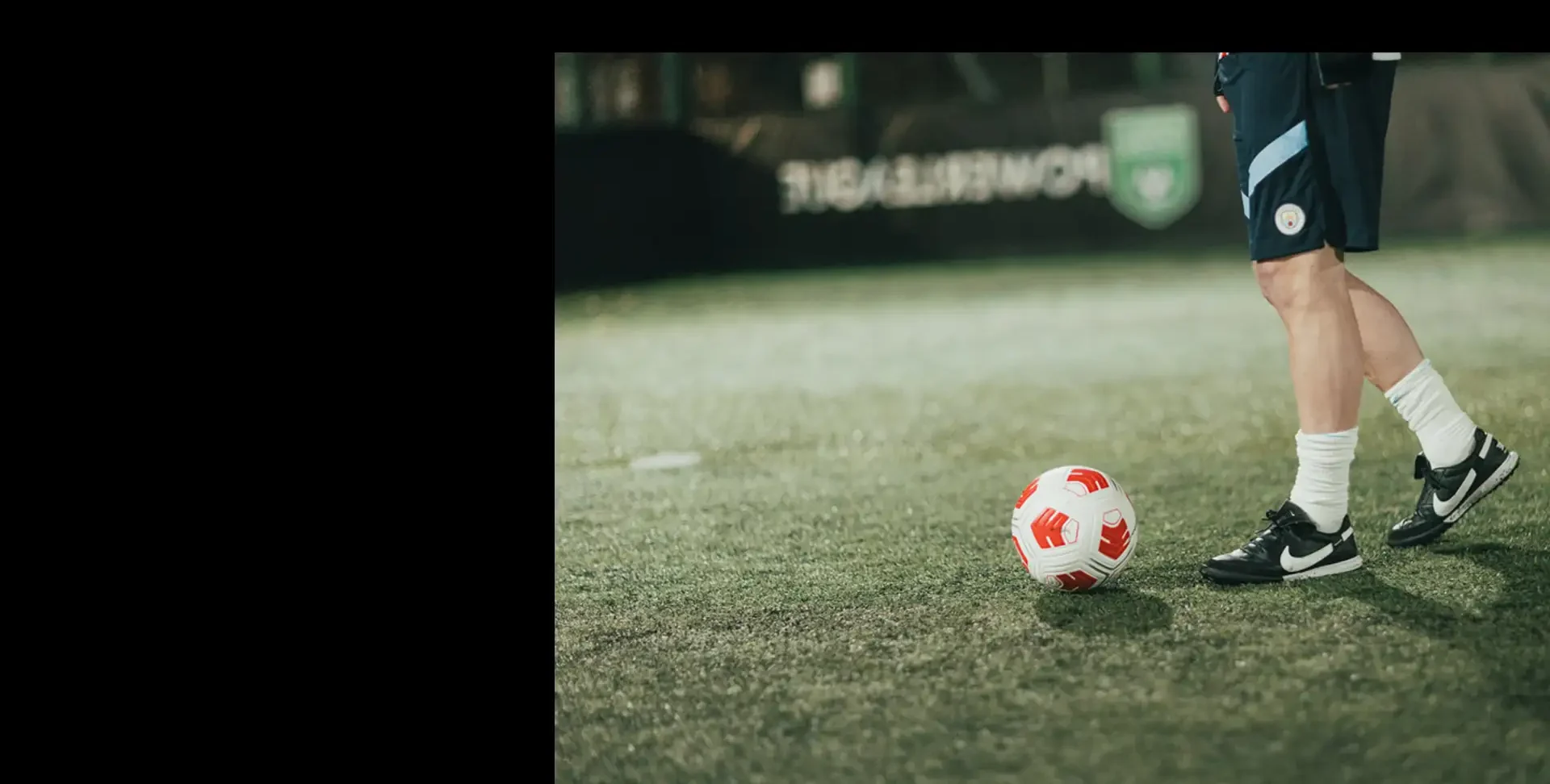 A person dressed in sportswear, including white socks and black Nike soccer shoes, is standing on a grass field near a red and white soccer ball. The setting appears to be a soccer field with a partially visible banner hinting at upcoming football tournaments in the background. © Powerleague