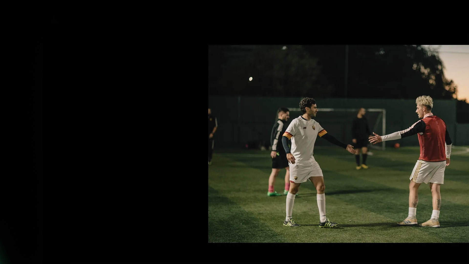A group of soccer players on a field at night. Two players, one in a white uniform and the other in a red jersey, are engaged in a fist bump. Other players stand in the background near the goals. The field is dimly lit, emphasizing the players' actions—true teamwork found at careers at Powerleague.