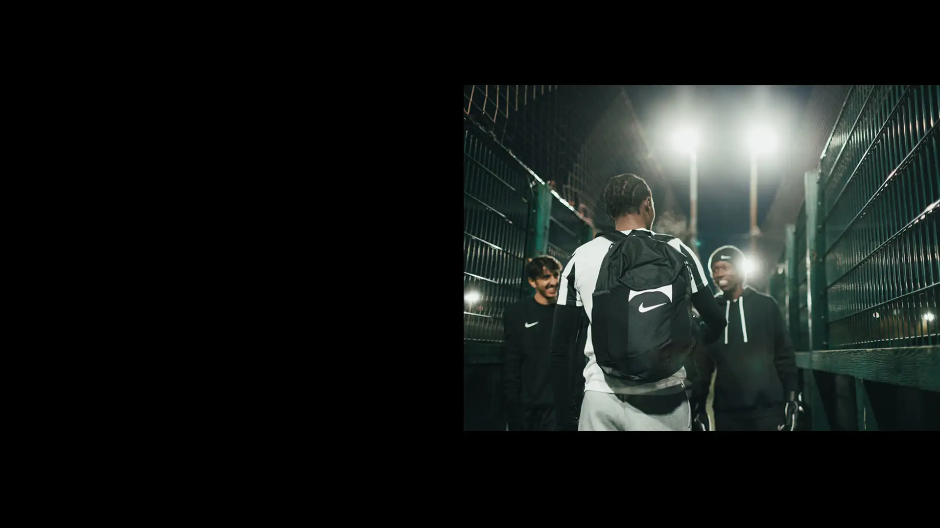 A night-time scene of three young men standing together outdoors near a fence. One person, wearing a black Nike backpack, has their back to the camera. The other two, possibly student athletes, are facing him and smiling in their dark athletic clothing under bright lights from the nearby 5-a-side field. © Powerleague