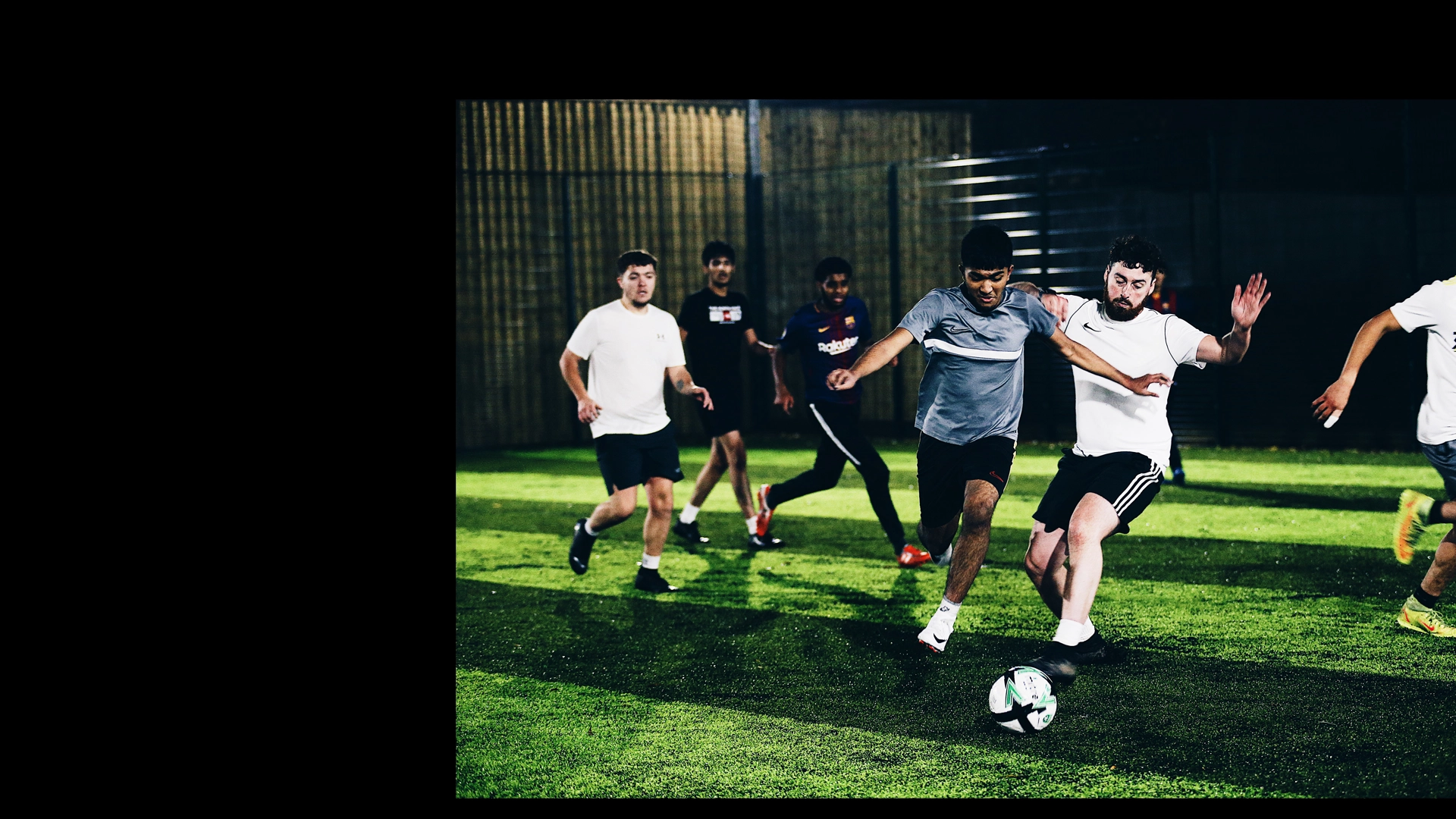A group of people are playing an intense game of indoor soccer on a green turf field, showcasing their health and stamina. One player in a white shirt and black shorts is attempting to kick the ball while another player in a gray shirt and black shorts is trying to block him. Others are in the background. © Powerleague