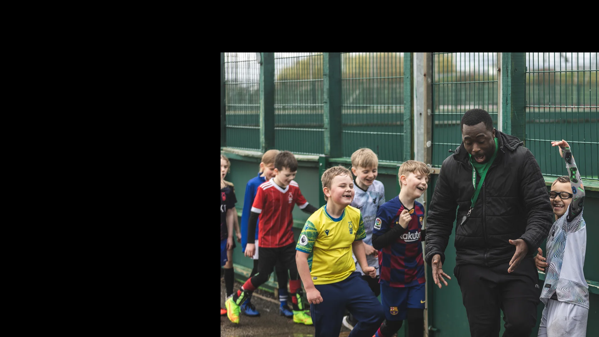 A group of young boys wearing various soccer uniforms enthusiastically follow a smiling man in a black jacket. They are outside near a fenced area, enjoying what appears to be a fun and energetic moment, reminiscent of the lively atmosphere at football holiday camps. © Powerleague