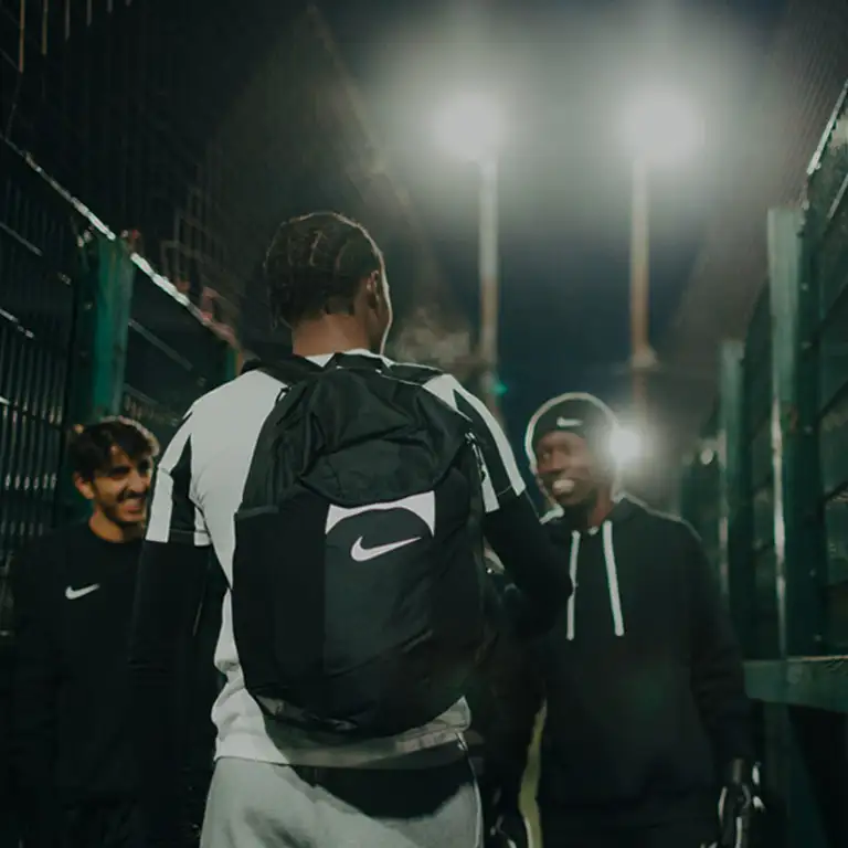 Three people are standing outdoors at night near a fence. The person in the foreground, possibly a student, has a backpack and is facing away from the camera. The other two are smiling and wearing athletic clothing, perhaps just finished with a 5-a-side game. Bright lights illuminate the background. © Powerleague