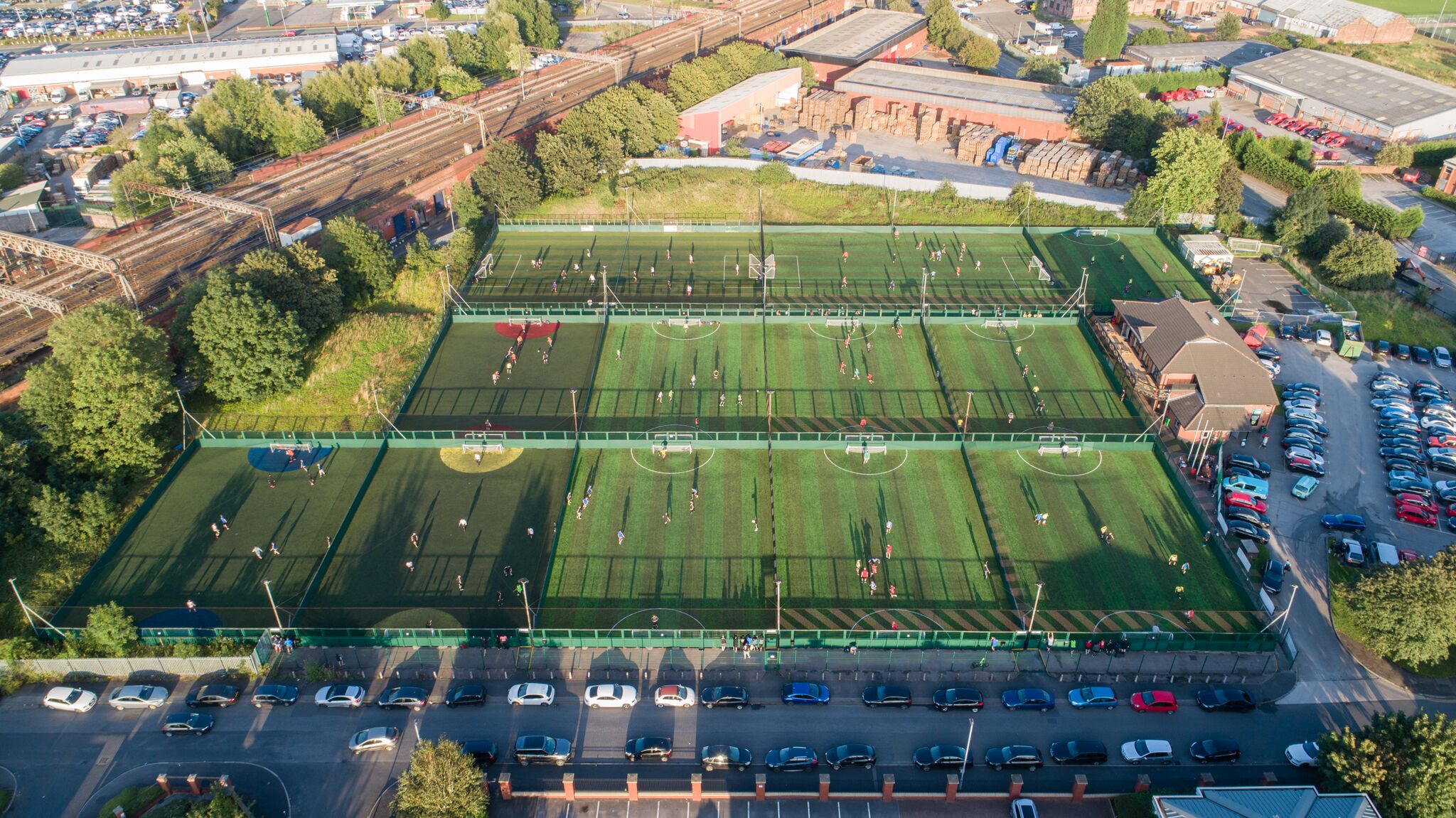 Aerial view of a sports complex featuring several adjacent artificial turf soccer fields buzzing with players from local football leagues. Surrounding the fields are parked cars, adjacent buildings, trees, and a bustling parking lot. The area is bathed in daylight. © Powerleague