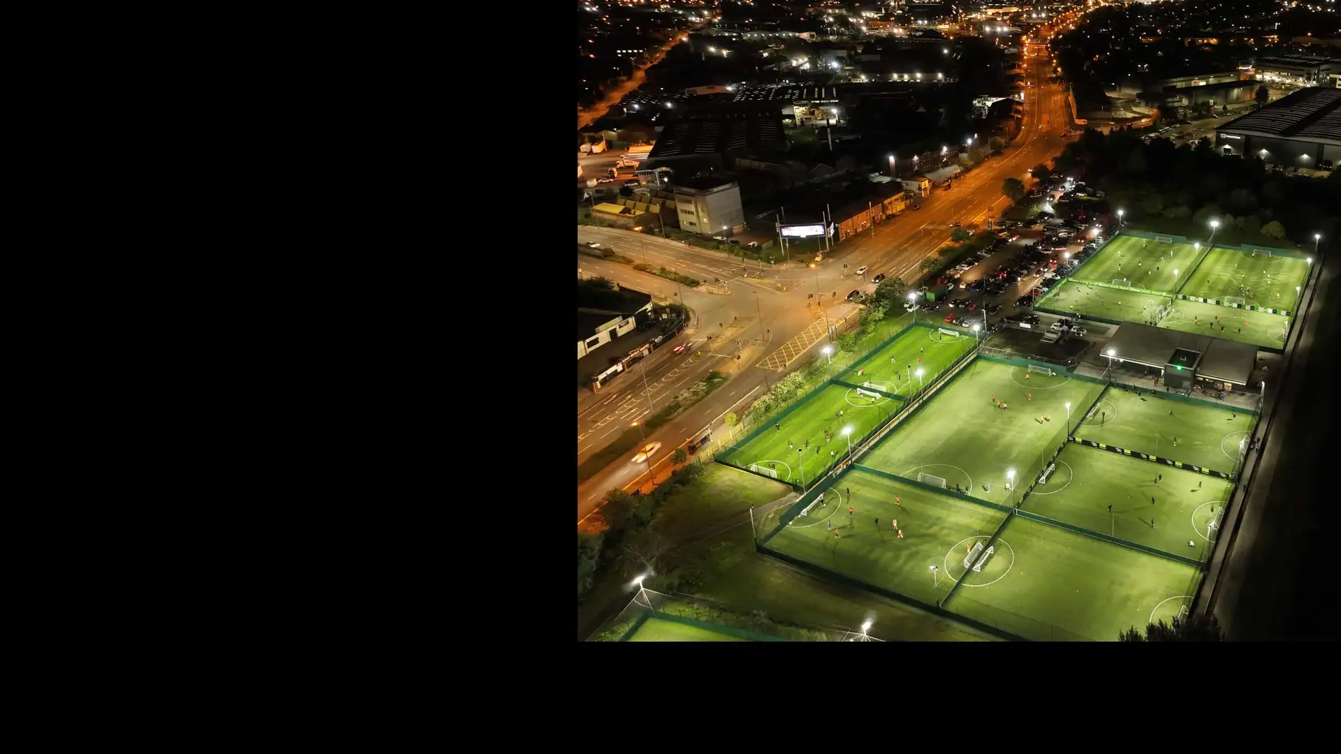 Aerial view of several brightly lit sports fields at night, surrounded by netted fences. Players from local football leagues can be seen engaging in various games on the fields. Nearby, city streets are illuminated by streetlights, with buildings and vehicles visible in the background. © Powerleague