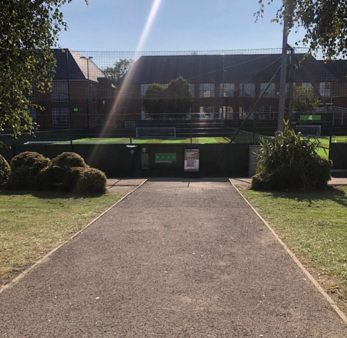 A path leads to a fenced-in sports field under a clear blue sky, where local football leagues compete. Brick buildings and trees stand in the background as sunlight casts a bright beam across the scene. © Powerleague