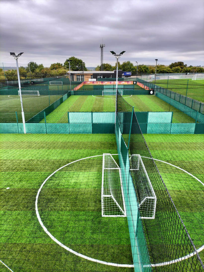 Aerial view of a fenced multi-purpose sports facility featuring artificial turf soccer fields, often bustling with local football leagues. Two small goals are placed on the nearest field. Surrounding trees and cloudy skies add a serene backdrop to the lively scene. © Powerleague