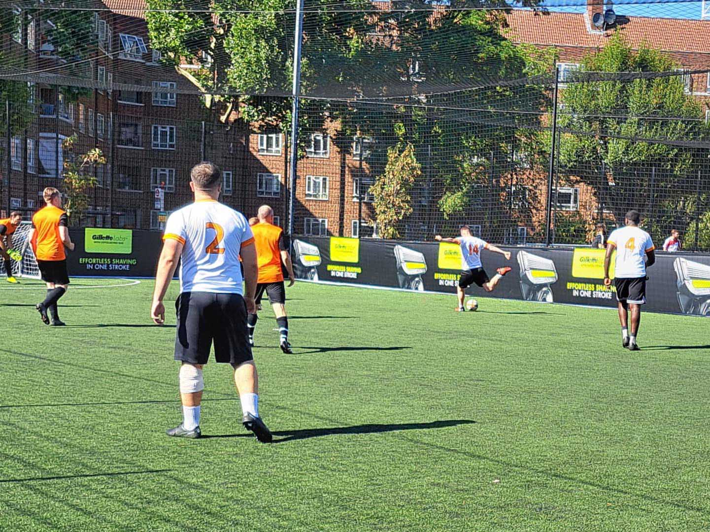 A group of men playing 5 a side football on a sunny day. Representing local football leagues, players in white and orange jerseys compete on a fenced field, surrounded by buildings and trees in the background. © Powerleague