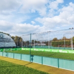 Outdoor football pitch with green turf and surrounding netting. A white dome labeled 
