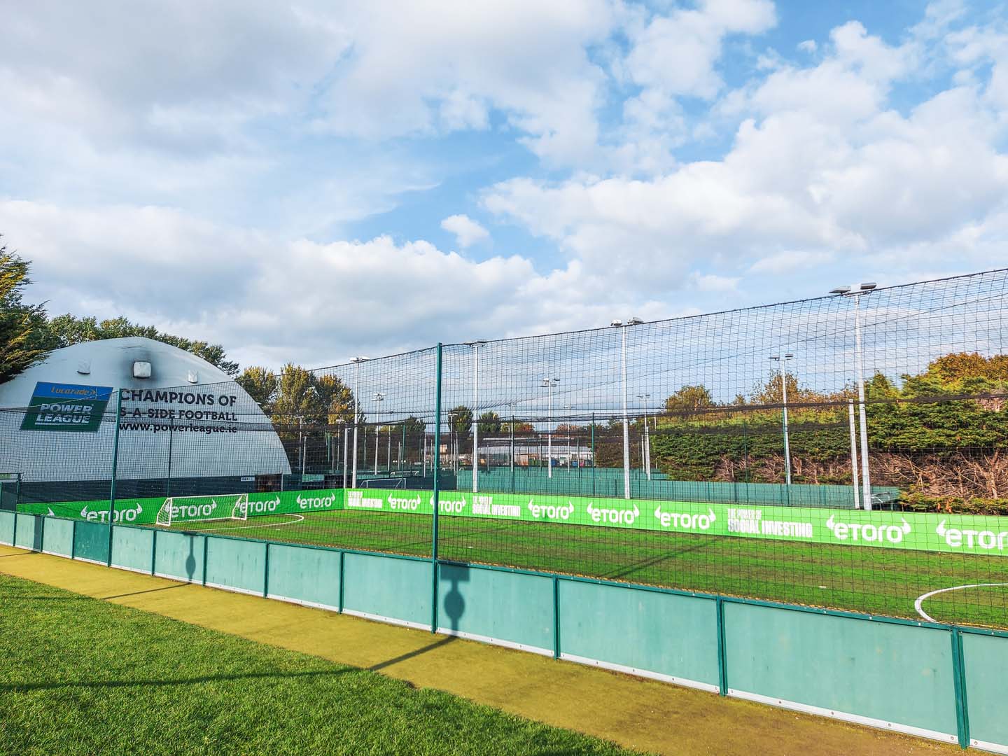 Outdoor football pitch with green turf and surrounding netting. A white dome labeled 