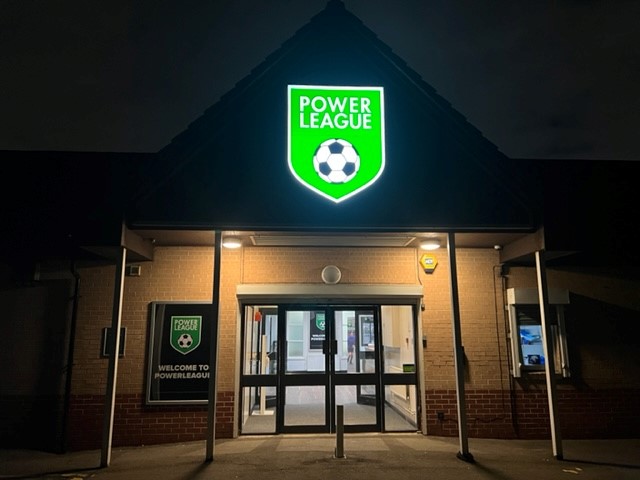 The entrance of the Power League building at night is illuminated by a bright green sign featuring a soccer ball emblem, symbolizing its ties to local football leagues. Through the glass doors, the inviting interior and a welcoming sign on the left wall beckon visitors inside. © Powerleague