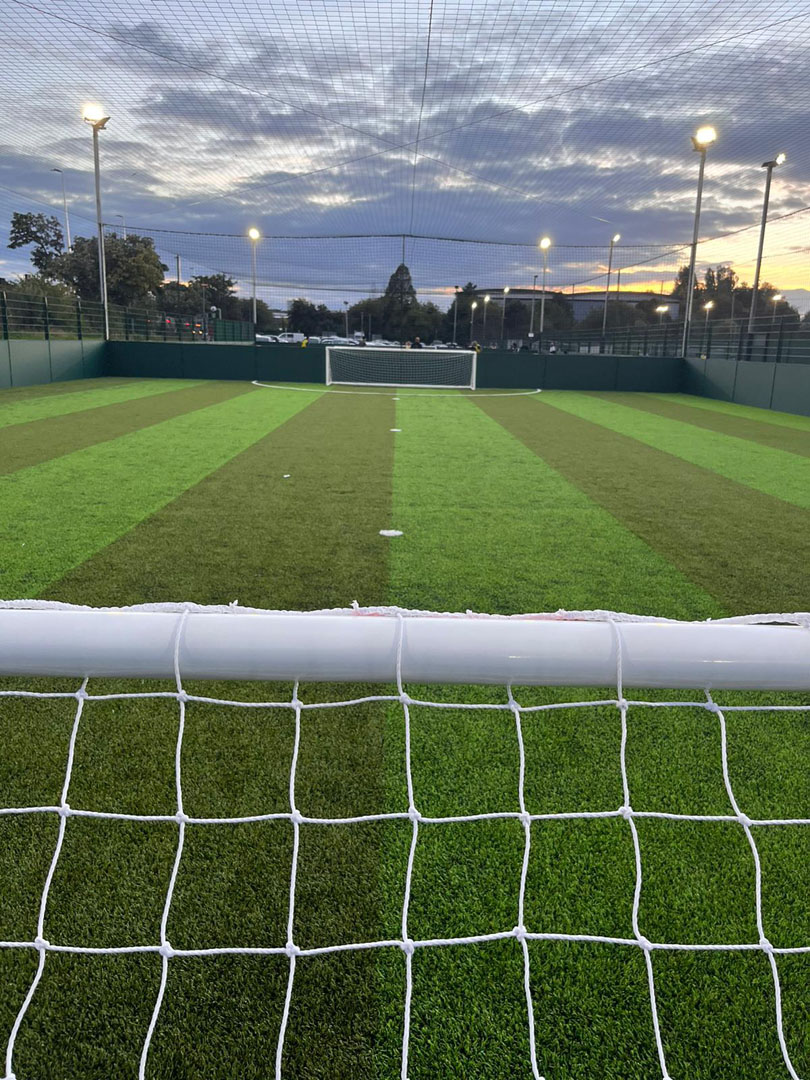 View of a soccer field from behind a goal, under a cloudy sky at dusk. The well-lit turf hosts local football leagues and is surrounded by a green fence. Trees and other urban elements are visible in the background, painting an idyllic scene for community sports enthusiasts. © Powerleague
