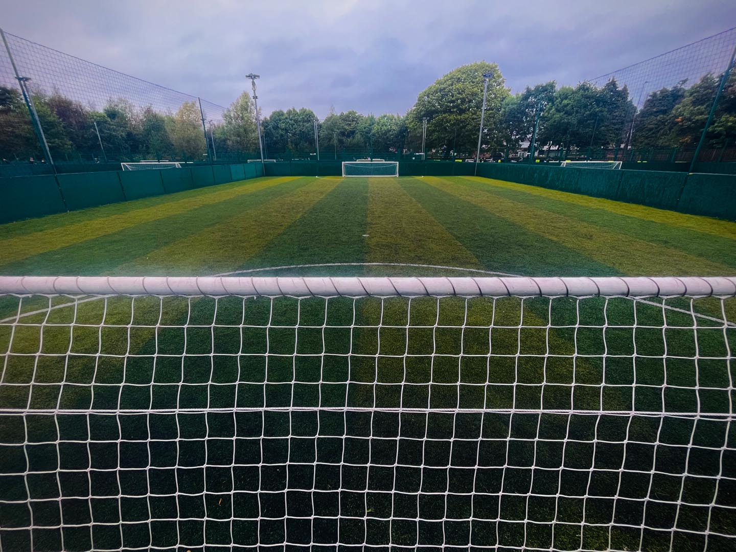 View of a lush green soccer field seen from behind a goal net, often used for local football leagues. The pitch is surrounded by a tall fence and bordered by trees, with another goal visible in the distance under a cloudy sky. © Powerleague