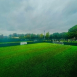 A small outdoor soccer field with green artificial turf is surrounded by a tall, green fence. Perfect for local football leagues, the field sits empty with a goalpost at one end. Trees and another field are visible under a cloudy sky in the background. © Powerleague