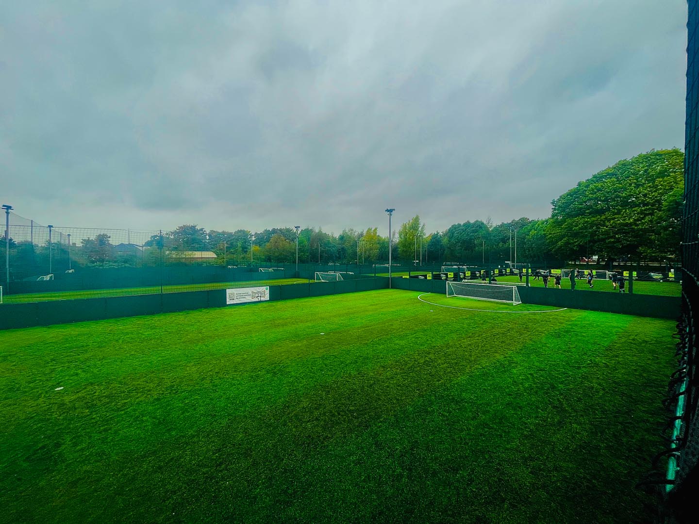 A small outdoor soccer field with green artificial turf is surrounded by a tall, green fence. Perfect for local football leagues, the field sits empty with a goalpost at one end. Trees and another field are visible under a cloudy sky in the background. © Powerleague