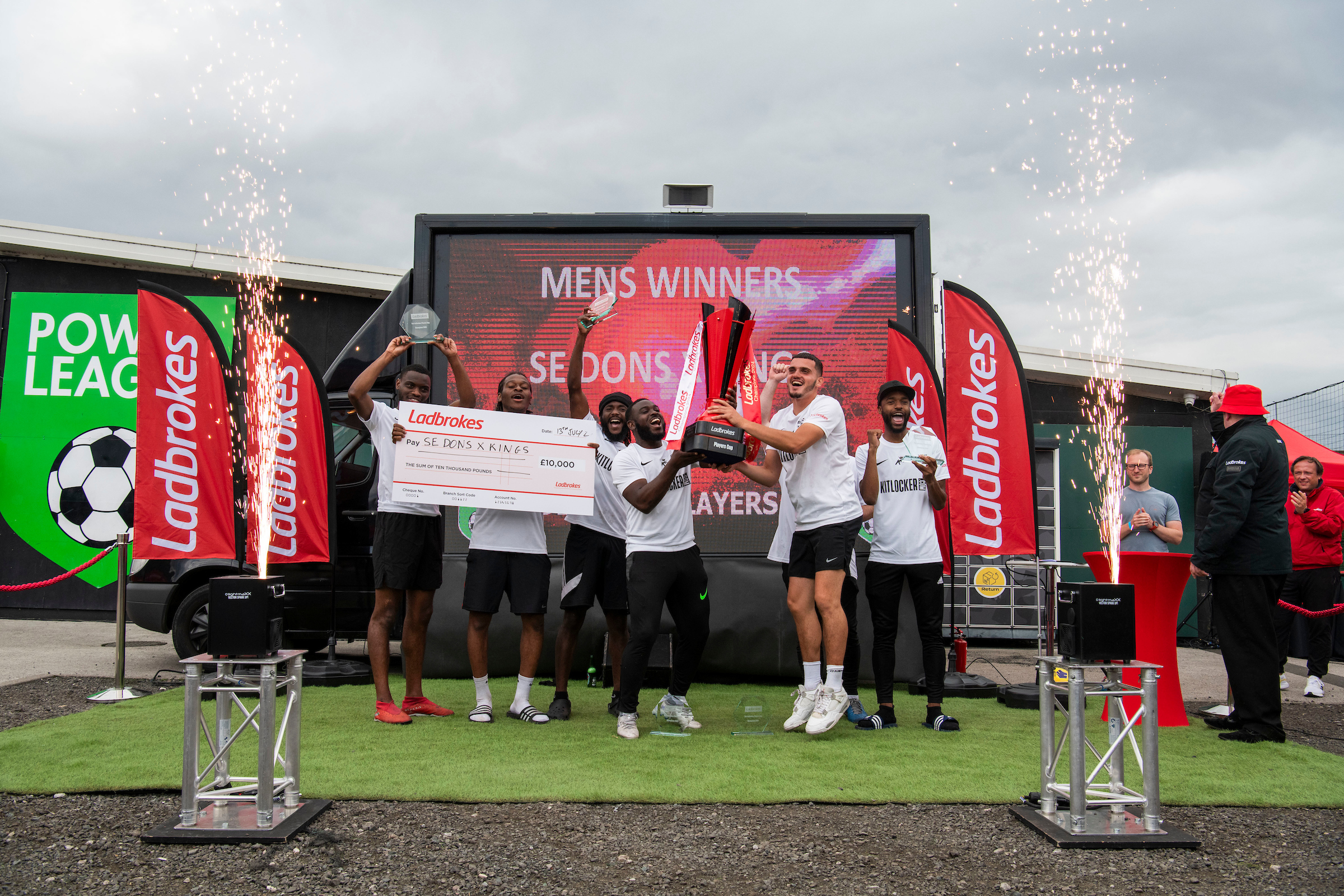 A team of six men, champions of the local football leagues, pose triumphantly with a trophy and a large check on stage. They stand in front of a screen displaying 