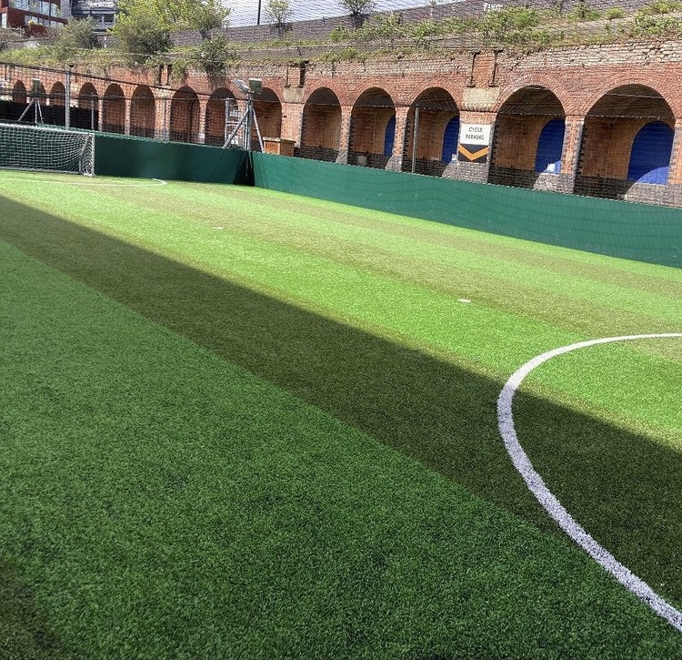 A small outdoor soccer field with artificial turf, ideal for local football leagues, is surrounded by green fencing. Brick arches and a sign with text are visible in the background as sunlight casts shadows across the field. © Powerleague