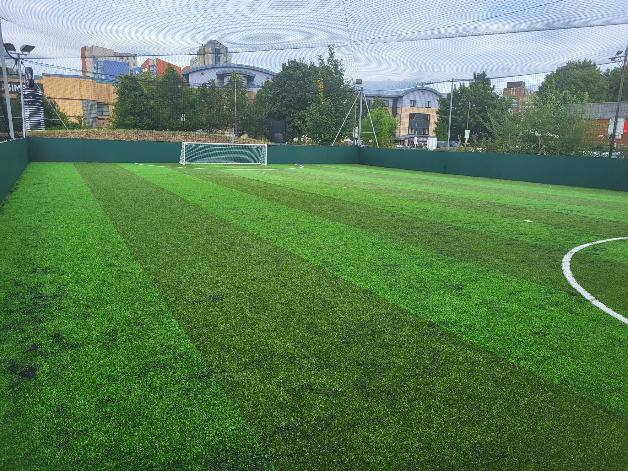 A well-maintained outdoor soccer field with artificial turf, featuring a goal at one end and often hosting local football leagues. The field is surrounded by high netting, with urban buildings visible in the background under a partly cloudy sky. © Powerleague
